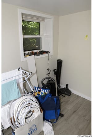 This is a photograph of a utility or storage room showing various household items and equipment stored haphazardly, including coiled cables, tools, a clothes drying rack, and a blue bag. The room has white walls, a window with a view of foliage, and 