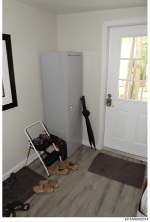 This is a photograph of an interior residential space showing an entryway or mudroom area. The room contains a white metal folding chair with various items on it, a tall gray storage cabinet, wooden sandals/shoes on the floor, an umbrella, and a brow