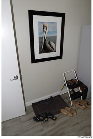 This is a photograph of an interior room space showing a framed pelican photograph hung on a white wall, with various personal items including shoes, a stepladder with clothing, and other belongings on the floor. The image appears to be evidence docu
