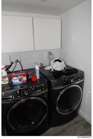 This is an evidence photograph showing a residential laundry room containing two black front-load washing machines, cleaning supplies, and miscellaneous household items. The photo appears to be part of property documentation in the investigation, cap