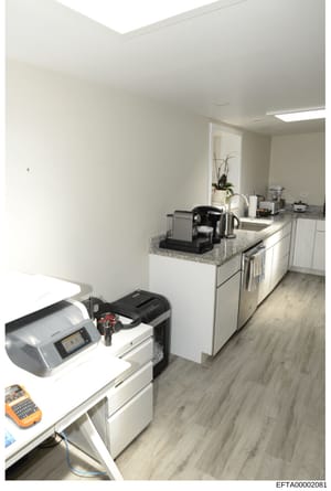 This photograph shows a modern kitchen space with contemporary cabinetry, granite countertops, and integrated appliances. The kitchen also contains office equipment including a label maker and other devices, suggesting this space served dual purposes