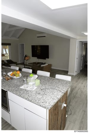 This is a photograph of a contemporary kitchen and adjoining living area, showing a granite-countertop island with white cabinetry, stainless steel appliances, and an open-concept layout with a media room visible in the background. The photo appears 