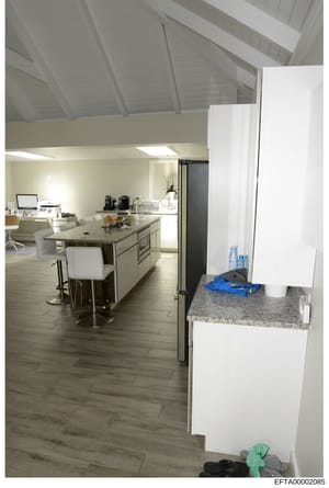 This is a photograph of a modern, open-plan kitchen and living area with contemporary furnishings and finishes. The image shows a large kitchen island with granite countertops, white cabinetry, stainless steel appliances, and vaulted ceilings with sk