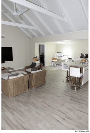 This is a photograph of a contemporary interior space featuring an open-plan layout with living and working areas. The space displays modern furnishings including wicker chairs, a kitchen counter, desk workspace, and high white ceilings with exposed 