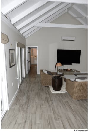 This is a photograph of an interior room with contemporary design features, including exposed white beam vaulting, wooden plank flooring, wicker furniture, and a wall-mounted flat-screen television. The image appears to be documentation of a property
