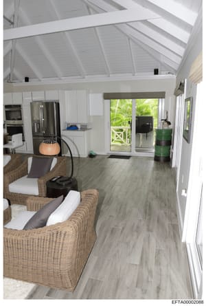 This is an evidence photograph showing the interior of a residential space, featuring a modern open-plan kitchen and living area with white cabinetry, stainless steel appliances, vaulted ceiling, and wicker furniture. The photograph documents the lay