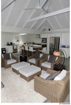 This is an interior photograph showing a modern, light-filled residential space with an open-concept design combining a kitchen and living area. The image depicts high ceilings with exposed white beams, stainless steel appliances, wicker seating furn