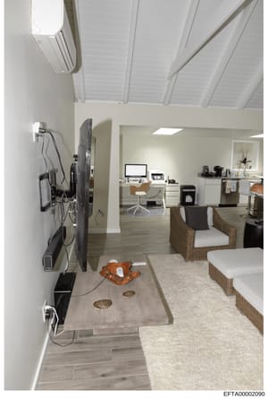 This is a photograph of a contemporary residential interior space showing an open-plan living area with an integrated home office. The room features modern furnishings including wicker lounge chairs, a flat-screen television mounted on the wall, a de