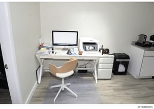 This is a photograph of a modern office or workspace interior, featuring a white desk setup with an iMac computer, printer, office supplies, and a wood-and-white ergonomic chair. The room has light gray walls, wood-grain flooring, and includes filing
