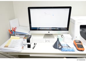 This is a photograph of a workspace showing a desk setup with an Apple iMac computer, papers, writing implements, and office supplies. The desk appears to be part of an investigation evidence documentation, with various documents and materials visibl