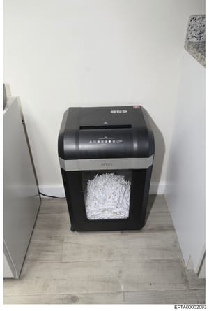 Photograph of a black and silver Ativa brand paper shredder containing shredded documents. The shredder is positioned on a light wood-tone floor against white walls. This image is documented evidence, likely taken during a search or seizure related t