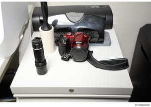 This is an evidence photograph showing a red Nikon digital camera with accessories, including a telephoto lens, lens cap, camera strap, and what appears to be a charging dock or scanner device. The items are displayed on a white surface under control