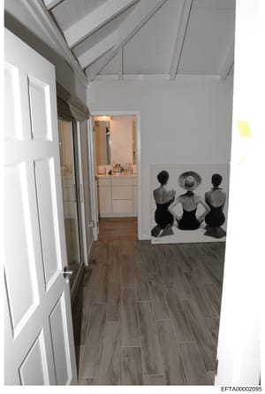 This photograph documents the interior of a residential property, showing a hallway with wood-grain flooring, white cabinetry, vaulted ceiling with skylights, and an open doorway revealing a bathroom vanity area. An artwork depicting three figures in
