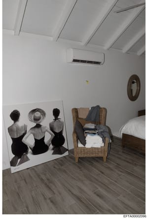 This is a photograph of a bedroom interior featuring a large artistic print depicting three female figures from behind, a wicker chair with folded clothing and textiles, a bed with white bedding, and modern amenities including an air conditioning uni