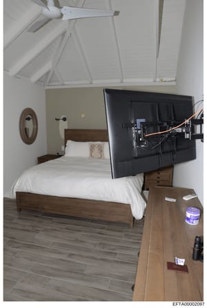 This photograph documents an interior bedroom space, featuring a wooden bed frame with white linens, a flat-screen television mounted on an articulating arm, and ceiling fixtures. The room appears to be photographed as part of property documentation 
