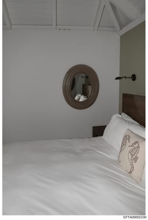 This is a photograph of a bedroom interior showing a made bed with white linens and decorative pillows, a round framed mirror on the wall, and a modern wall sconce. The image appears to be documentation of property interior details, possibly from one