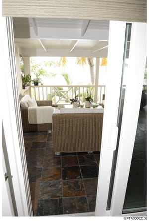 This is a photograph of a luxurious covered outdoor living space, showing a screened or covered porch with high-end wicker furniture, potted plants, and views to tropical landscaping. The image appears to be from an interior design or property docume