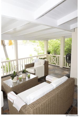 This is a photograph of an elegant covered outdoor space featuring wicker furniture with white cushions, potted plants, and views of tropical landscaping. The image shows a well-maintained residential porch or veranda with decorative white railings, 