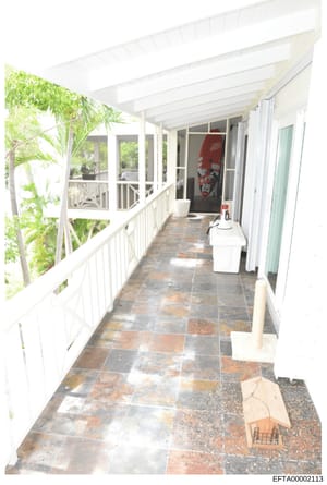 This is a photograph of a covered porch or corridor with white painted columns and railings, slate tile flooring, and large windows. The space appears to be part of a residential property with tropical landscaping visible through the windows. A red s