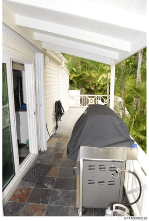 This is an evidence photograph showing an exterior covered patio or veranda area of a residential property. The image depicts a screened/covered outdoor space with slate flooring, white trim, tropical plants visible in the background, and various ite