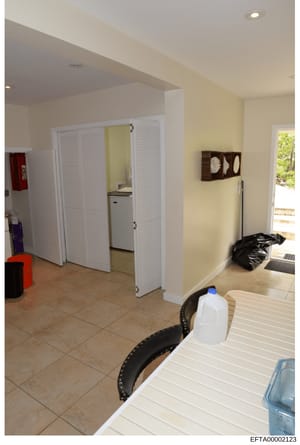 This is an evidence photograph showing the interior of a residential space, featuring a kitchen area with white louvered doors, yellow walls, tile flooring, and various household items and storage containers. The image appears to be documentation of 