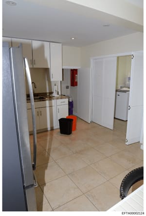 This is a photograph of a residential kitchen interior, showing appliances, cabinetry, flooring, and various household items. The image appears to be part of property documentation or evidence collection related to the investigation, with a Bates num