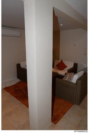 This photograph documents an interior room space featuring a central white structural column dividing the area, with seating furniture on one side including wicker chairs with cushions and a red decorative pillow. The room has light-colored walls, re