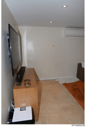 This is an evidence photograph showing the interior of a room, likely from one of Epstein's properties. The image depicts a minimally furnished space with a flat-screen television mounted on a wooden console, recessed ceiling lights, air conditioning