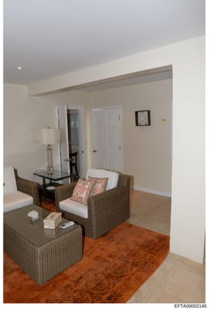 This is a photograph of a residential interior room featuring contemporary furnishings. The space shows a seating area with wicker furniture, neutral décor, and minimal personal items. The photo appears to be documentation of property interior condit