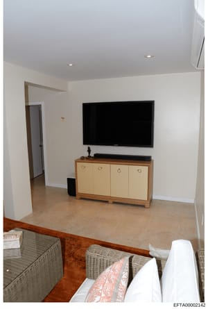 This is a photograph of an interior room featuring a flat-screen television mounted on a white wall above a wooden cabinet with light-colored doors. The room has tile flooring, recessed lighting, and wicker seating visible in the foreground. The imag
