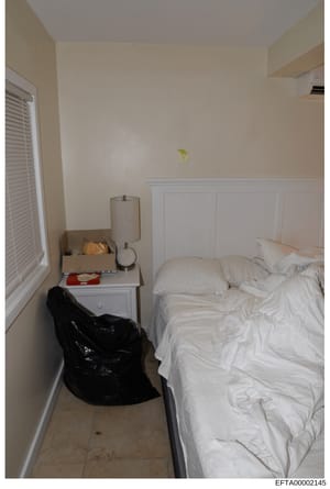 This is a photograph of a bedroom interior showing a bed with white linens, a bedside table with a lamp and various items, a black bean bag chair, venetian blinds on a window, and cream-colored walls with white wainscoting. The photo appears to be ev