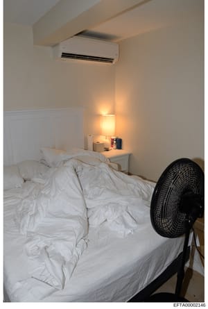 This is a photograph of a bedroom interior showing a bed with white linens, a nightstand with a lamp, and an air conditioning unit mounted on the wall. The image appears to be documentary evidence from a property seizure or inventory, likely taken du