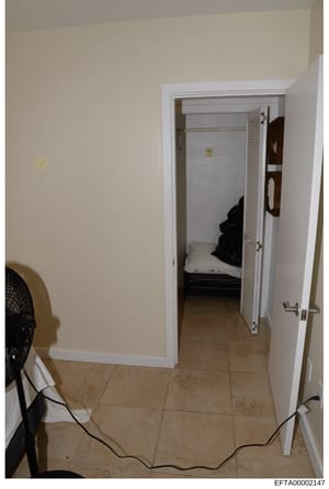 This is an evidence photograph showing an interior room with beige walls, tan tile flooring, and an open closet door revealing a bedroom beyond. A black electric fan is visible on the left side, and electrical cords run across the floor. The photogra