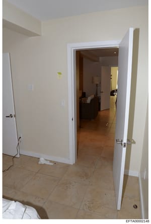 This is a photograph of an interior residential space showing a doorway with white trim leading into an adjacent room with dark wood flooring. The image captures beige/cream-colored walls, recessed shelving on the left, electrical outlets, and a view