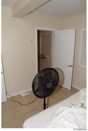 This is an evidence photograph showing the interior of a room with cream-colored walls, tile flooring, and minimal furnishings. A black pedestal fan is prominently positioned in the center of the room, with an open doorway visible in the background a
