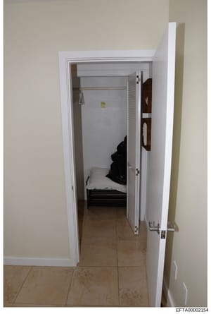 This is a photograph of a small interior room or closet space showing a bed with black bedding and white pillow against the far wall. The space appears narrow with white-painted walls and tile flooring. The image is dated and catalogued as evidence f
