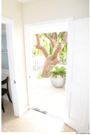 This is an evidence photograph showing the interior of a residential space featuring a glass door or sliding door opening to an outdoor area. The image captures cream-colored walls, white trim, tile flooring, and a view of tropical landscaping includ