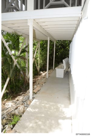 This photograph depicts a white-columned covered walkway or passageway with a concrete floor, bordered by tropical vegetation including palm trees. The structure appears to be part of a larger property and features white overhead beams and what appea