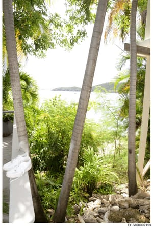 This is a photograph of a tropical oceanfront property showing a white structure (likely a building or deck), lush vegetation including palm trees, and a clear view of the ocean with an island or peninsular landform visible in the distance. The image