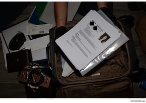 This is a photograph of seized evidence showing documents, travel tickets, and personal items contained within a brown suitcase or travel bag. A white document with a photo ID visible is prominently displayed, along with what appears to be airline ti