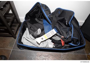 This is a forensic evidence photograph showing a black and blue suitcase or travel bag containing various clothing items, including what appears to be undergarments and other fabrics. A white label with a barcode is visible on one of the items inside