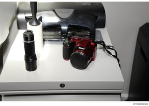 This is an evidence photograph showing a red Nikon digital camera, a black telephoto lens, and what appears to be a printer or scanner device arranged on a white surface. The photo documents seized or collected equipment, likely relevant to the inves