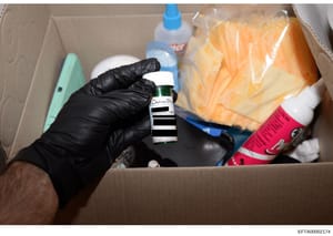 This is an evidence photograph showing a black-gloved investigator's hand holding a small glass vial with a white cap and striped label reading 'Chelsea Dyer' inside a cardboard box containing various household items. The photo documents items seized