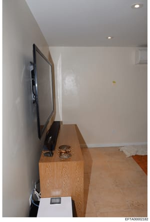 This is a photograph of an interior room showing a wall-mounted flat-screen television on a wooden stand/console, along with decorative items and furnishings. The image appears to be evidence documentation from a property associated with the investig