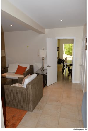 This is a photograph of a modern interior room, likely a bedroom or suite, showing contemporary furnishings and design. The image depicts woven rattan chairs with white cushions and an orange accent pillow, a side table with lamp, white walls with ex