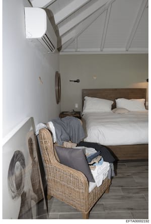 This is a photograph of a bedroom interior, likely from one of Jeffrey Epstein's properties, showing a made bed with white linens, wooden headboard, and various furnishings including a wicker chair with clothing and linens, wall-mounted decorative it