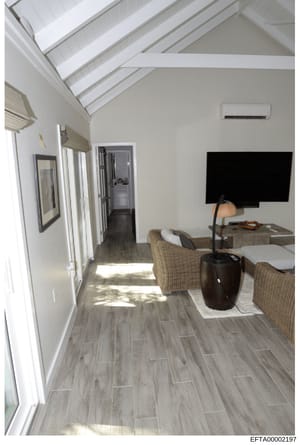 This photograph shows the interior of a modern residential room, featuring contemporary design elements including vaulted ceilings with skylights, light wood-look flooring, wicker furnishings, and a wall-mounted flat-screen television. The image appe
