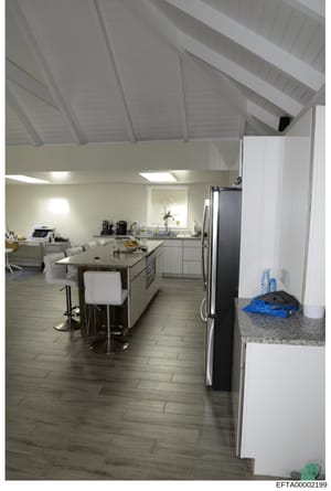 This photograph shows a modern kitchen and connected living area, featuring contemporary design elements including white cabinetry, stainless steel appliances, an island with seating, and vaulted ceilings. The image appears to be documentation of a r