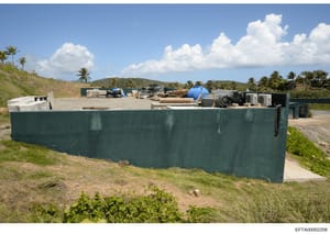 This photograph shows an outdoor construction or storage site enclosed by a large teal-colored metal fence. The facility contains various building materials, equipment, and supplies including wooden pipes, a blue water tank, metal containers, and HVA