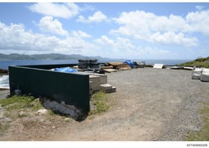 Photograph of a waterfront storage or work area showing various maritime equipment, construction materials, and storage structures along a rocky coastline. The image depicts a property with ocean views and mountains in the background, containing orga